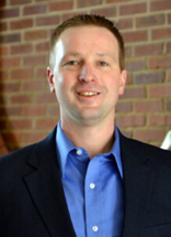 Smiling professional in navy blazer and blue shirt against brick wall