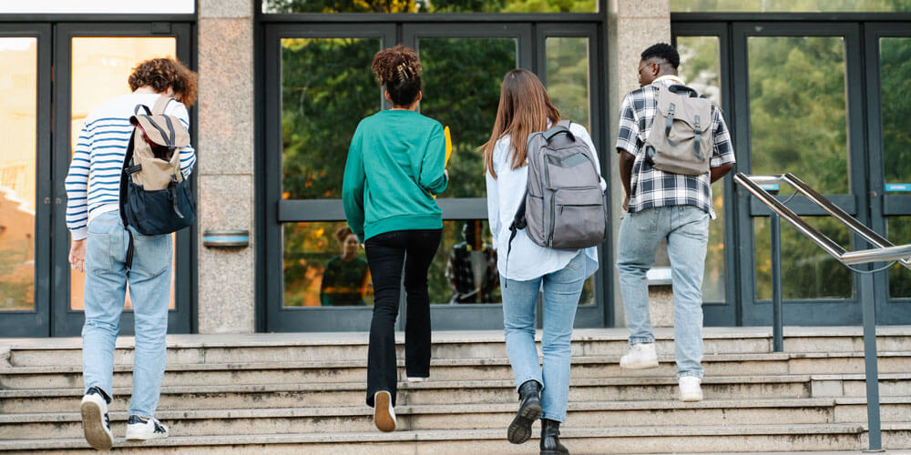 Four students with backpacks walking up concrete stairs near glass building entrance