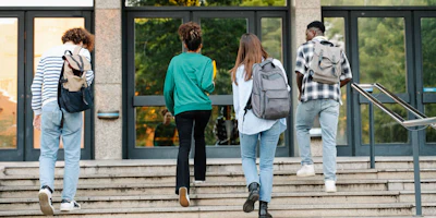 Four students with backpacks walking up concrete stairs near glass building entrance