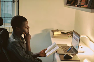 Man in plaid shirt studying documents at desk with laptop in bright office space