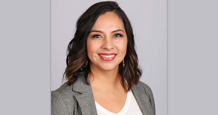 Professional headshot of a woman with dark wavy hair, red lipstick, wearing a gray blazer over white top, smiling against neutral gray background