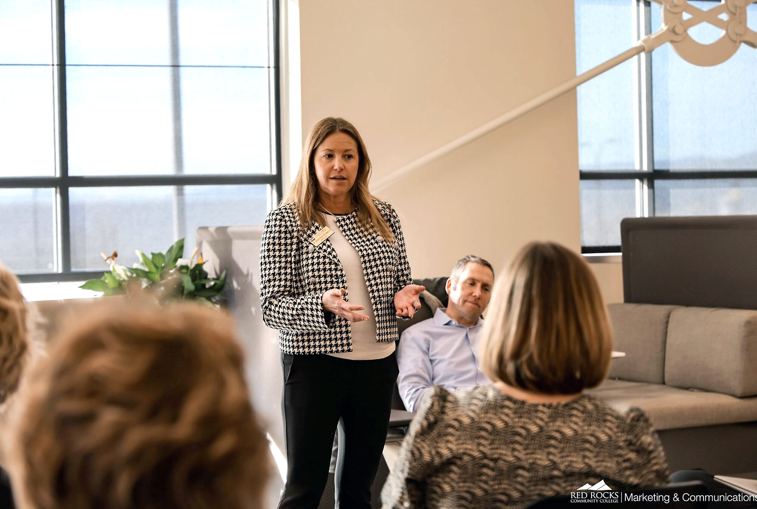 Professional woman presenting to colleagues in modern office with floor-to-ceiling windows and contemporary decor