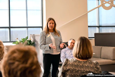 Professional woman presenting to colleagues in modern office with floor-to-ceiling windows and contemporary decor
