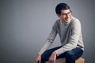Young man wearing glasses and gray sweater smiling while sitting on wooden stool against gray background