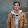 Young man smiling at camera wearing tan jacket and light blue hoodie against textured wall
