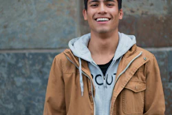 Young man smiling at camera wearing tan jacket and light blue hoodie against textured wall