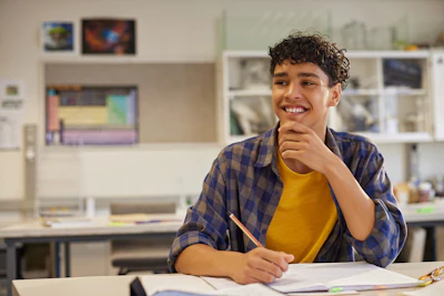 Smiling student in plaid shirt and yellow shirt at desk in classroom with shelving and computer monitors in background