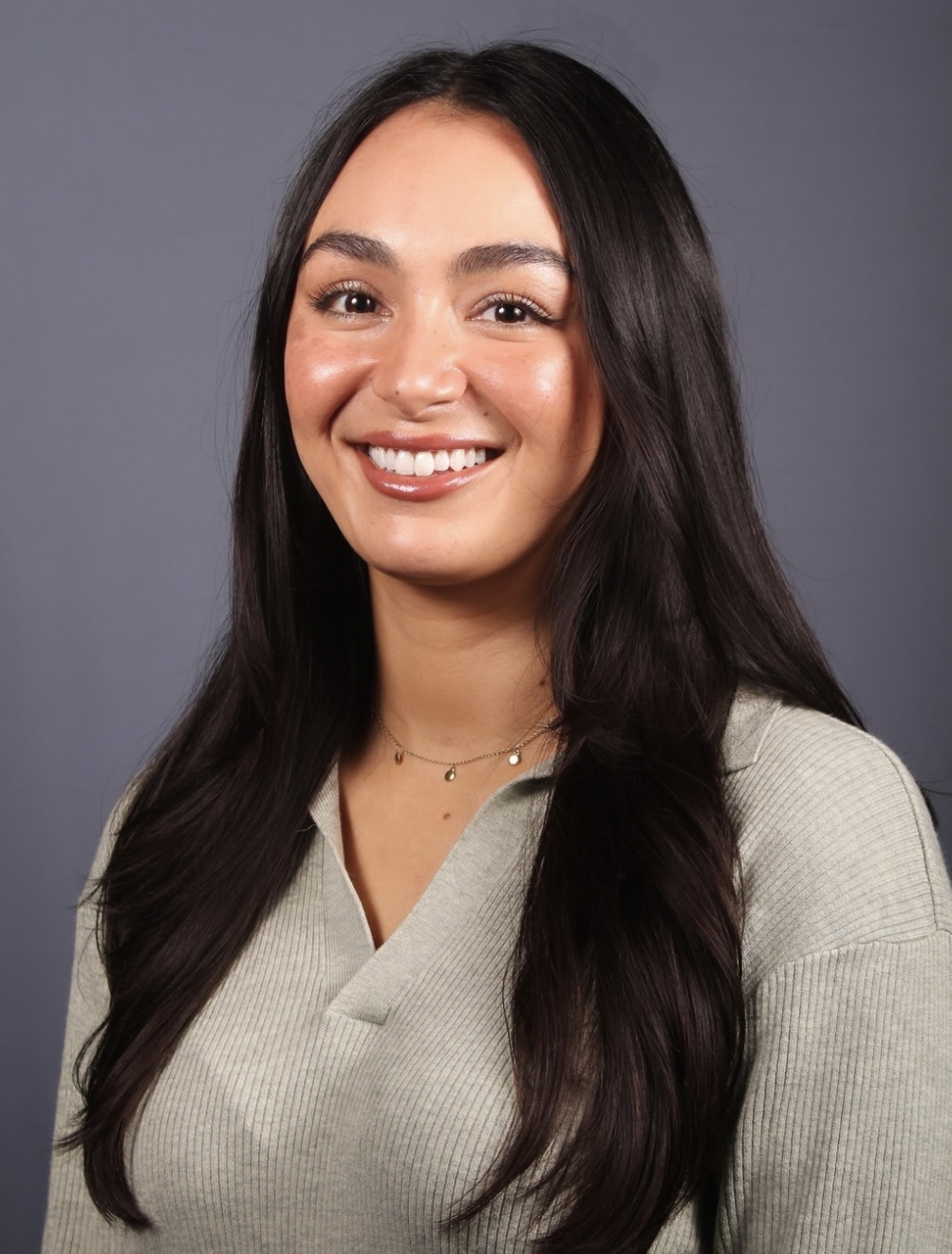 Professional headshot of a smiling woman with long dark hair wearing a light gray ribbed sweater and delicate necklace against a gray background